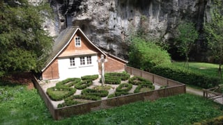 Blick auf ein kleines Häuschen mit eingezäuntem Garten vor Felswand. 