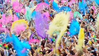 In einer Menschenmenge werfen die Leute gelbe, viollete, rosa, gelbe und blaue Pulver in die Luft.