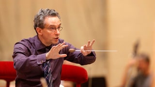 Ein Dirigent mit grauem Haar und Brille.