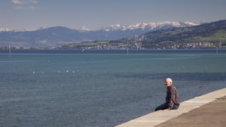 Ein älterer Mann sitzt am Ufer des Bodensees, in der Ferne die schneebedeckten Berge.