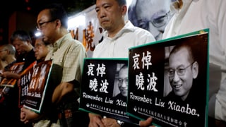 Männer stehen in einer Reihe, sie halten Plakate mit Fotos von Liu und chinesischem Text in ihren Händen.