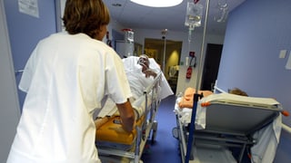 Eine Pflegefachfrau stösst ein Bett mit einem Patienten durch einen Spitalkorridor. Eine Pflegefachfrau stösst ein Bett mit einem Patienten durch einen Spitalkorridor.
