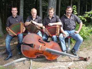 Die vier Musiker sitzen am Waldrand auf einer Bank. Vor ihnen liegt ein Kontrabass am Boden. Drei der Musiker halten ein Schwyzerörgeli auf den Knien.