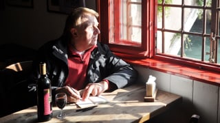 Hanns-Josef Ortheil sitzt mit einem Glas Wein und Notitzbuch an einem Tisch und schaut aus dem Fenster.