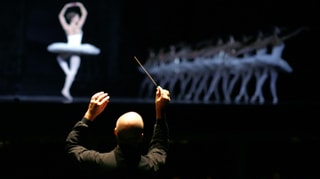 Ein Dirigent von hinten fotografiert, auf der Bühen Belletttänzerinnen.