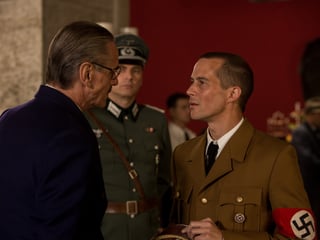 Filmstill: Ein Mann im dunklen Anzug unterhält sich mit einem Mann in Nazi-Uniform.