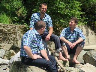 Drei junge Männer in karierten Hemden sitzen barfuss auf Steinen an einem Flussufer.