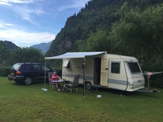 Wohnwagen mit aufgeklapptem Vordach, darunter sitzt eine Frau. Weiter hinten steht ein Auto mit holländischem Kontrollschild. Tessiner Berglandschaft.