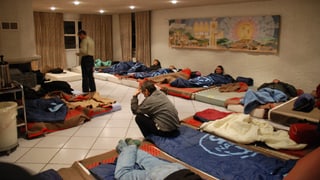 Junge Männer liegen auf Betten am Boden in einem grossen Raum.