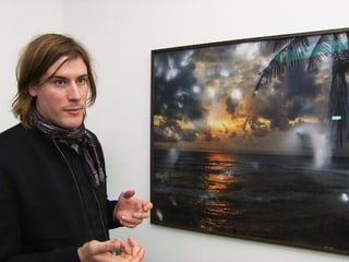 Julian Charrière vor einer Fotografie eines Strandes im Sonnenuntergang mit seltsam aufgehellten Flecken darin.