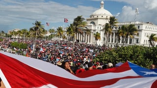 Man sieht im Vordergrund eine Fahne Puerto Ricos und im Hintergrund hunderte Demonstranten.