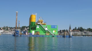 Ein gelbes Metallsilo steht auf einem Ponton im See. Dahinter die Silhoutte von Luzern. Ein gelbes Metallsilo steht auf einem Ponton im See. Dahinter die Silhoutte von Luzern.