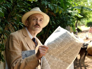 Mann mit Hut und einer Zeitung vor einem üpiigen, exotischen Wald.