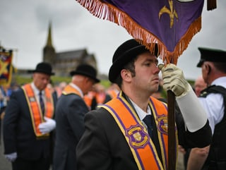 Im Vordergrund ein Umzugsteilnehmer mit einer Fahnenstange in der Hand. Daneben ein Polizist, im Hintergrund die Kirche von Drumcree.