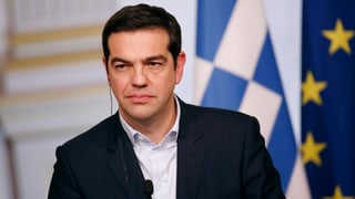 Tsipras vor einem Mikrofon, hinter ihm eine griechische und eine EU-Fahne.