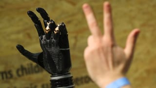 Eine Menschenhand zeigt einer Roboterhand, drei Finger in die Höhe zu strecken.