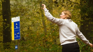 Zu sehen ist SRF 3 Moderatorin Kathrin Hönegger beim Joggen im Wald. Sie hält ein Smartphone in der Hand, ein Kopfhörer-Stöpsel ist im rechten Ohr, der zweite Ohrstöpsel ist rausgefallen. Links im Bild ist eine Vita-Parcours Tafel zu sehen.