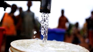 Aus einem Wasserhahn fliesst klares Wasser in einem alten Eimer. Im Hintergrund stehen afrikanische Menschen.