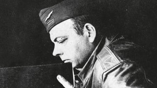 Schwarzeweissaufnahme: Saint-Eyupéry in Militärpiloten-Uniform hält ein grosses Buch.