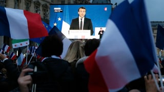 Emmanuel Macron auf einer Leinwand, davor sind Menschen mit Frankreich-Fahnen zu sehen.