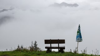 Eine Sitzbank und ein zugeklappter Sonnenschirm vor trüber Aussicht auf das verregnete und nebelverhangene Bergpanorama 