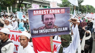 Muslime mit weissen Kappen tragen ein Transparent mit einem Foto von Basuki und der Aufschrift: «Verhaftet ihn, weil er den Islam beleidigt hat.»