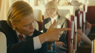 Junge mit blonden Haaren schiesst mit einem Finger in einem Schulsbus sitzend.