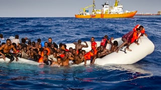 Afrikaner auf einem Gummiboot, das schon halb unter Wasser steht. Hinten ein Schiff.