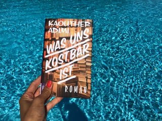 Annette König hält das Buch von Kaouther Adimi: «Was uns kostbar ist» vor blaues Wasser
