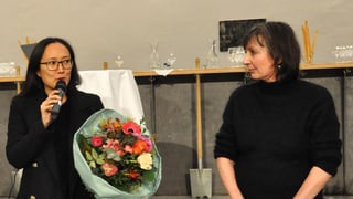 Eine Frau mit Blumenstrauss, in ein Mikrofon sprechend, neben ihr eine zweite Frau, ebenfalls stehend. Eine Frau mit Blumenstrauss, in ein Mikrofon sprechend, neben ihr eine zweite Frau, ebenfalls stehend.