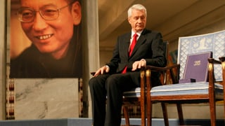 Ein Mann sitzt auf einem Stuhl, der Stuhl nebendran ist leer, auf der anderen Seite eine grosse Fotografie von Liu.