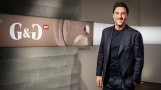 Mann mit kurzen dunklen Haaren, Brille und dunkelblauem Anzug vor einer Betonwand mit der Aufschrift «G&G» Mann mit kurzen dunklen Haaren, Brille und dunkelblauem Anzug vor einer Betonwand mit der Aufschrift «G&G»