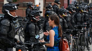 Eine Journalistin mit Mikrofon steht vor einer langen Reihe von Polizisten in schwarzer Kampfmontur.
