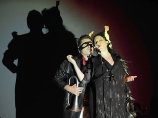 Eine Frau und ein Mann auf der Bühne. Er trägt eine Trompete und eine schwarze Maske. Sie ein schwarzes Federkleid. Eine Frau und ein Mann auf der Bühne. Er trägt eine Trompete und eine schwarze Maske. Sie ein schwarzes Federkleid.