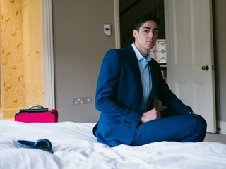 Ein junger Mann in blauem Anzug sitzt auf einem Bett.