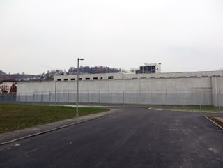 Eine asphaltierte Strasse führt zu einer hohen Betonmauer. Davor ein Zaun. Hinter der Mauer sieht man noch knapp den obersten Stock eines Gebäudes.