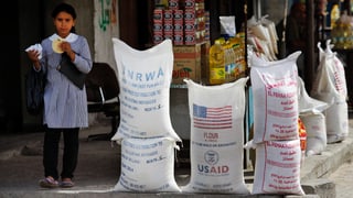 Ein Mädchen mit einem Brot in der Hand steht neben weissen Lebensmittelsäcken mit der Aufschrift «UNRWA» und «USAID».