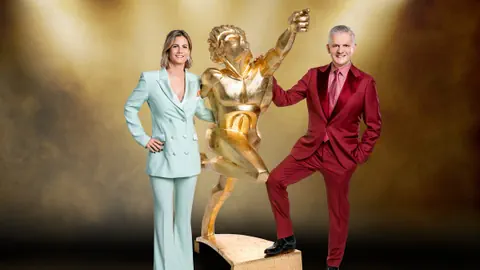 Das Moderatoren-Duo Fabienne Gyr und Rainer M. Salzgeber und in der Mitte eine Statue.