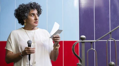 Person mit lockigem Haar hält Mikrofon und Papier vor bunter Wand.