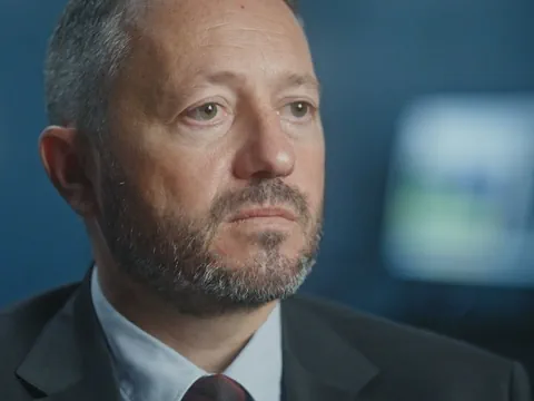 Mann mit Bart in Anzug vor unscharfem Hintergrund.