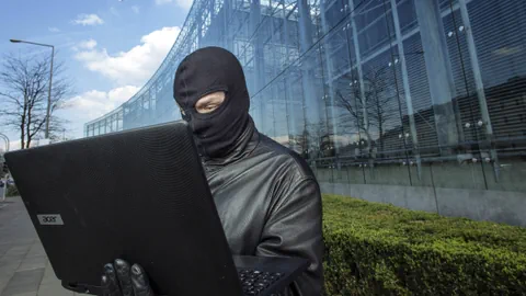 Person mit Sturmhaube arbeitet an Laptop vor Glasgebäude.