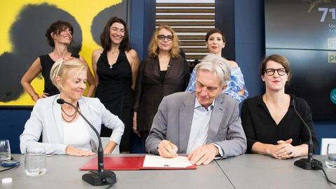Ein Mann unterschreibt ein Dokument. Links und rechts von ihm sitzen zwei Frauen.