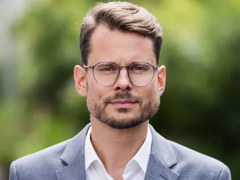 Mann mit Brille im Anzug vor unscharfem Hintergrund.