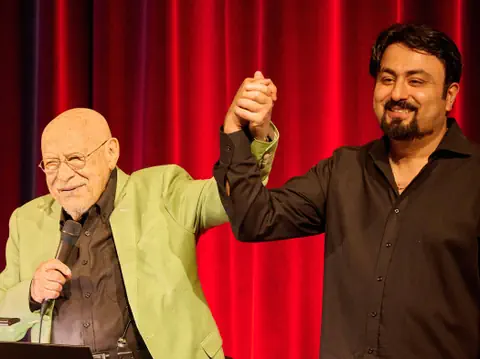 Zwei Männer halten Hände auf einer Bühne vor rotem Vorhang.