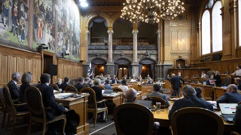 Menschen in einem historischen Sitzungssaal mit Wandgemälde und Kronleuchter.