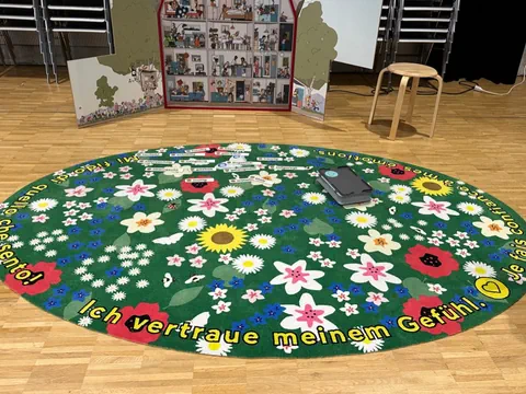 Runder Teppich mit bunten Blumen und deutschem Text, auf Holzboden, mit Stuhl und Dekoration im Hintergrund.