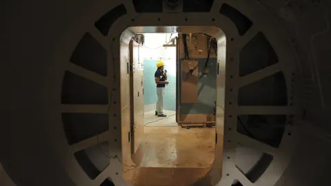 Person mit gelbem Helm in technischem Raum gesehen durch runde Türöffnung.