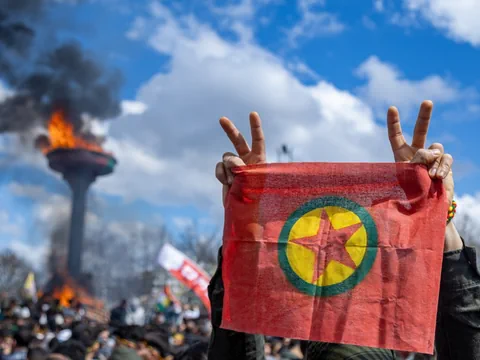 Person hält Flagge mit Symbol, davor grosse brennende Fackel unter bewölktem Himmel.