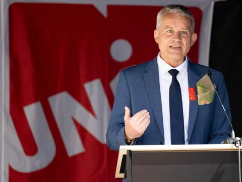 Mann hält Rede vor rotem Banner mit Mikrofon.