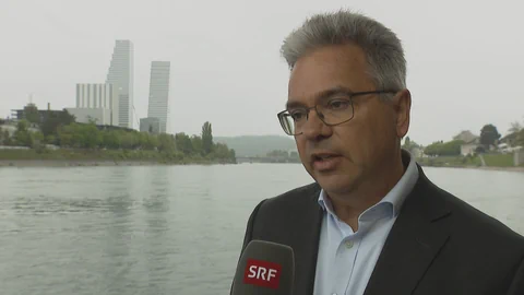 Mann am Fluss mit Mikrofon, Stadt im Hintergrund.